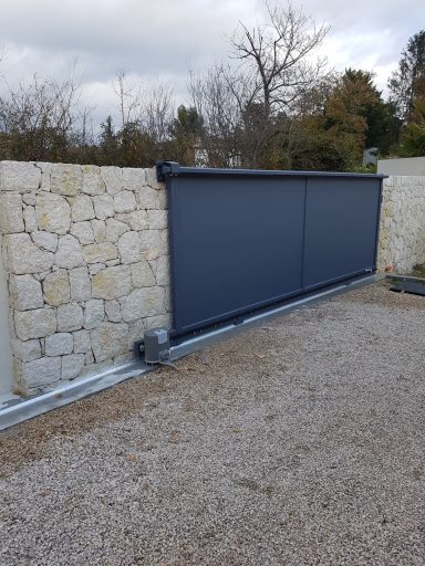 Portail coulissant gris en métal, entouré d'un mur en pierre, sur un chemin gravillonné.