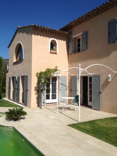 Maison avec fenêtres grises, patio, et piscine verte, sous un ciel bleu.